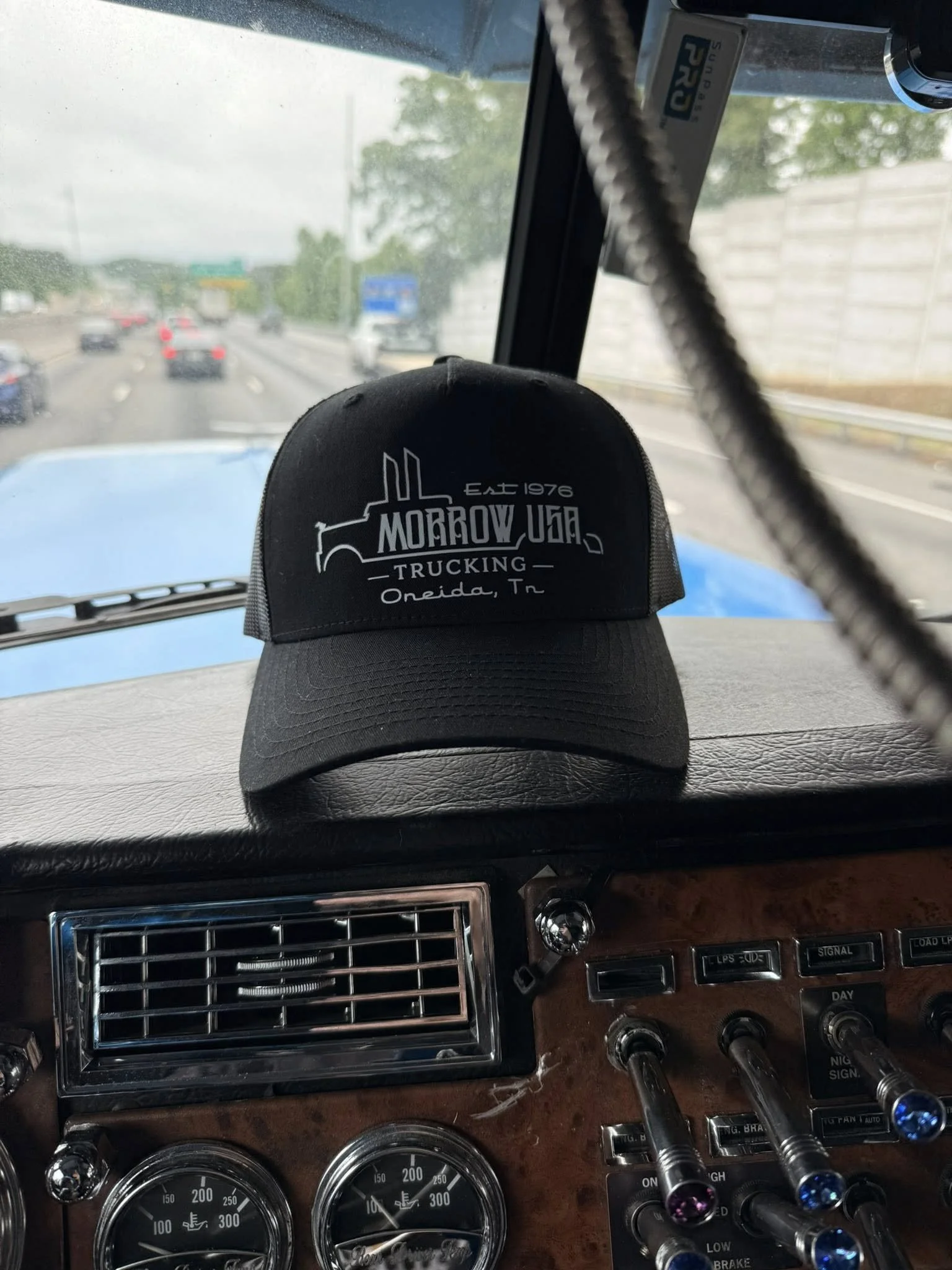The image shows the interior of a truck with a black cap placed on the dashboard. The cap has the text "MORROW UGA TRUCKING Oneida, Tn" embroidered on it. The vehicle is on a highway with visible traffic and a concrete wall on the right side. The view through the windshield shows multiple cars and a cloudy sky.