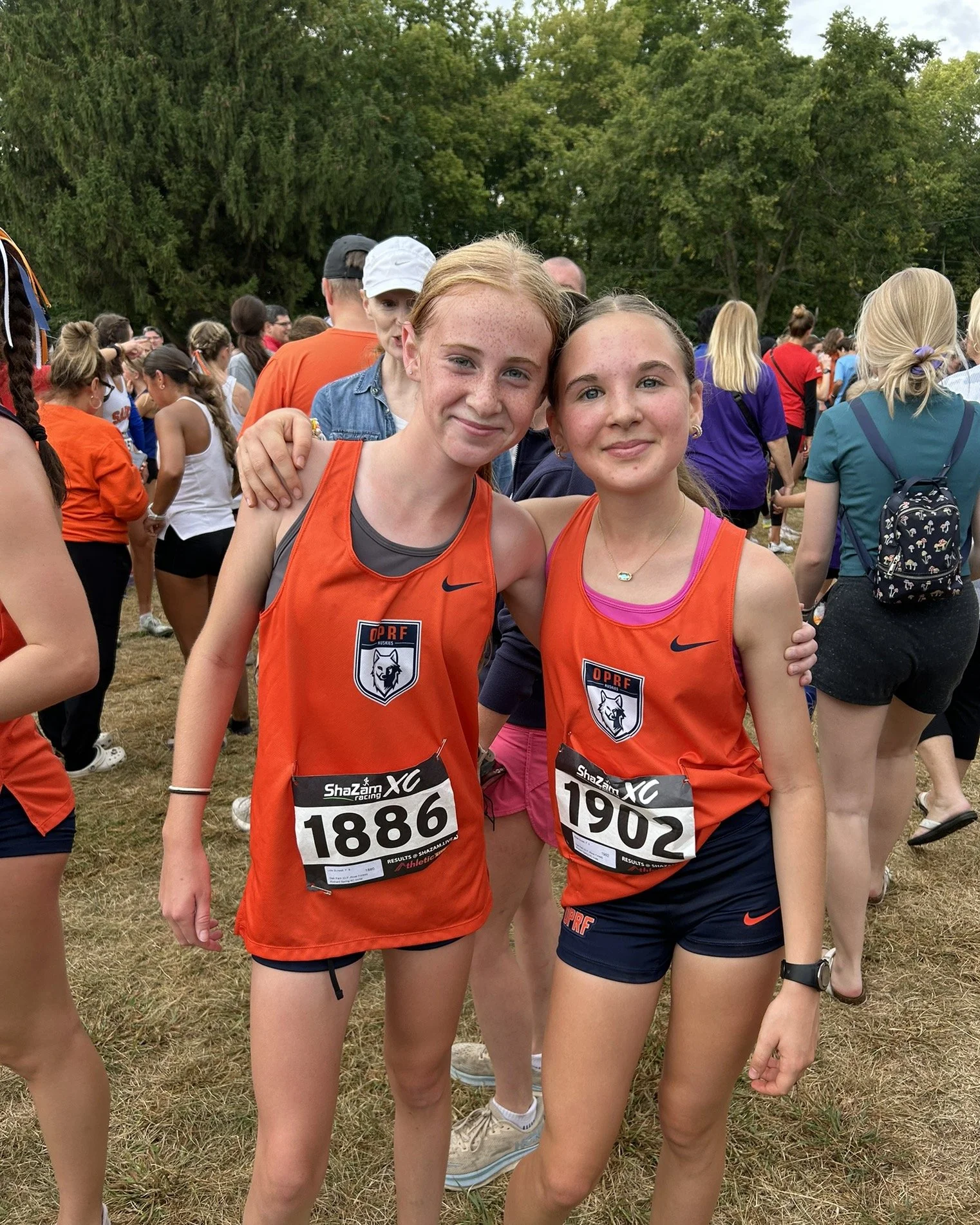 Two young women with light skin and freckles, wearing orange athletic Oak Park and River Forest tank tops with race bibs, standing arm in arm at an outdoor cross country race event surrounded by a crowd of people.