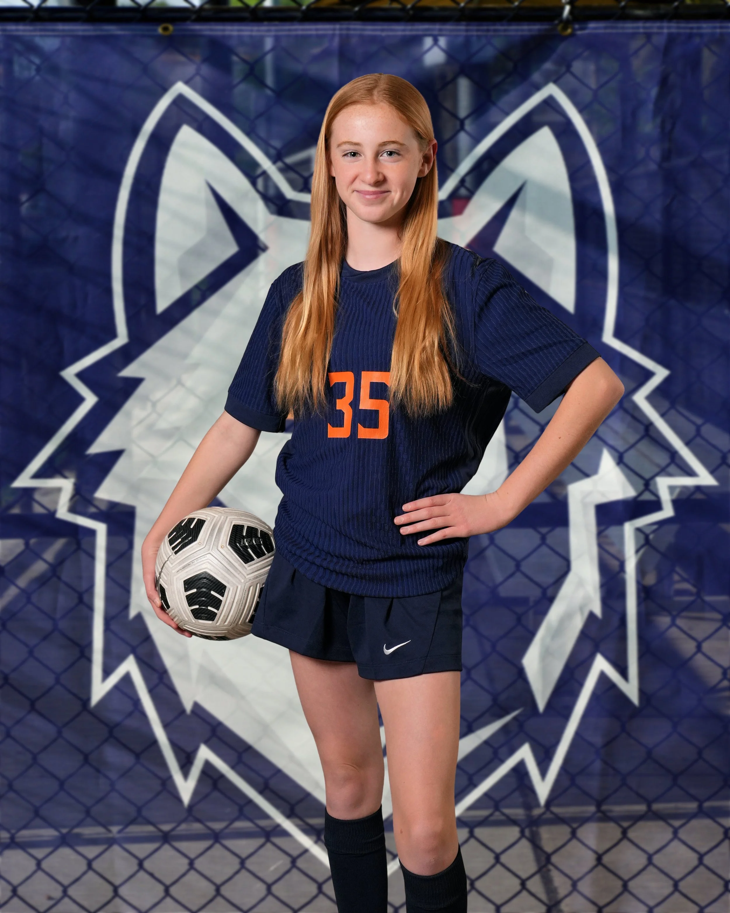 A young female soccer player with red hair, wearing a navy blue jersey with orange number 35 and black shorts, poses with a soccer ball in front of a blue and white banner with a star logo, standing on a soccer field.
