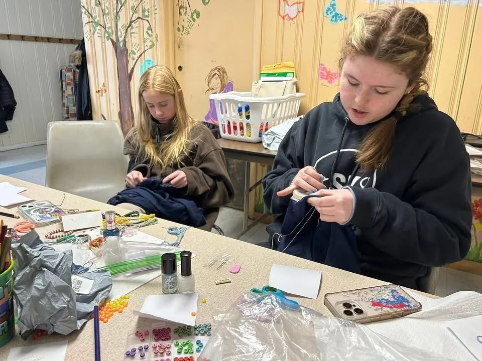 Two girls are sitting at a craft table, working on projects with various craft supplies like beads, thread, and decoration items, in a room with wooden walls and colorful decorations.