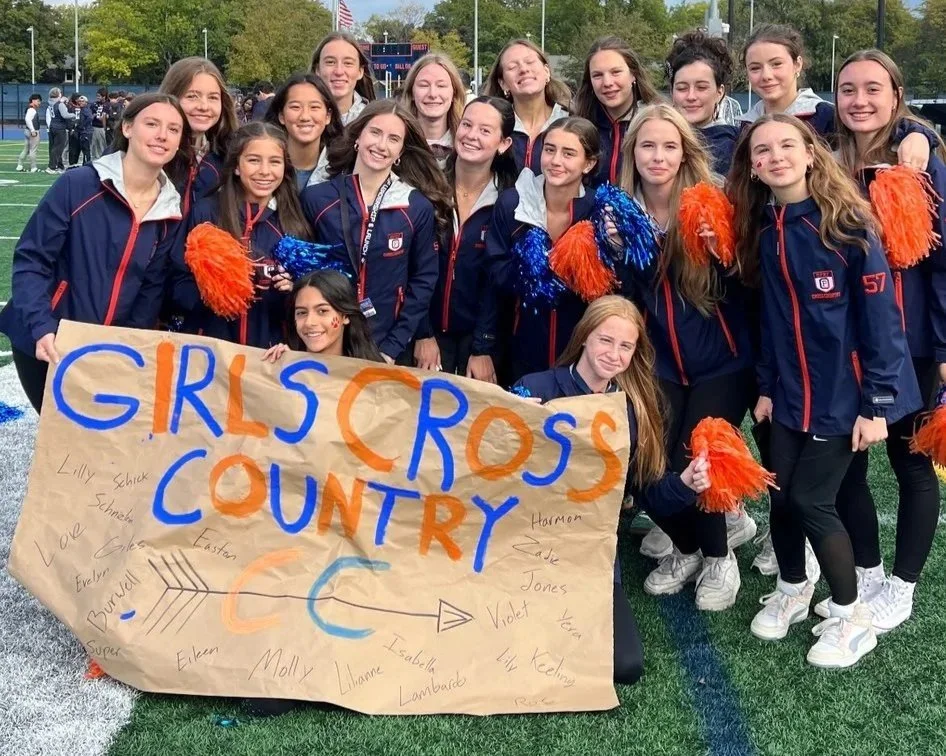 Group of young girls in warm ups holding pom-poms, smiling, standing on a sports field, holding a large handmade sign that reads 'Girls Cross Country CC' with various signatures.