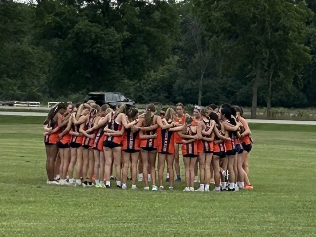 A group of young female cross country runners in matching orange and navy uniforms huddle together on a grassy field.