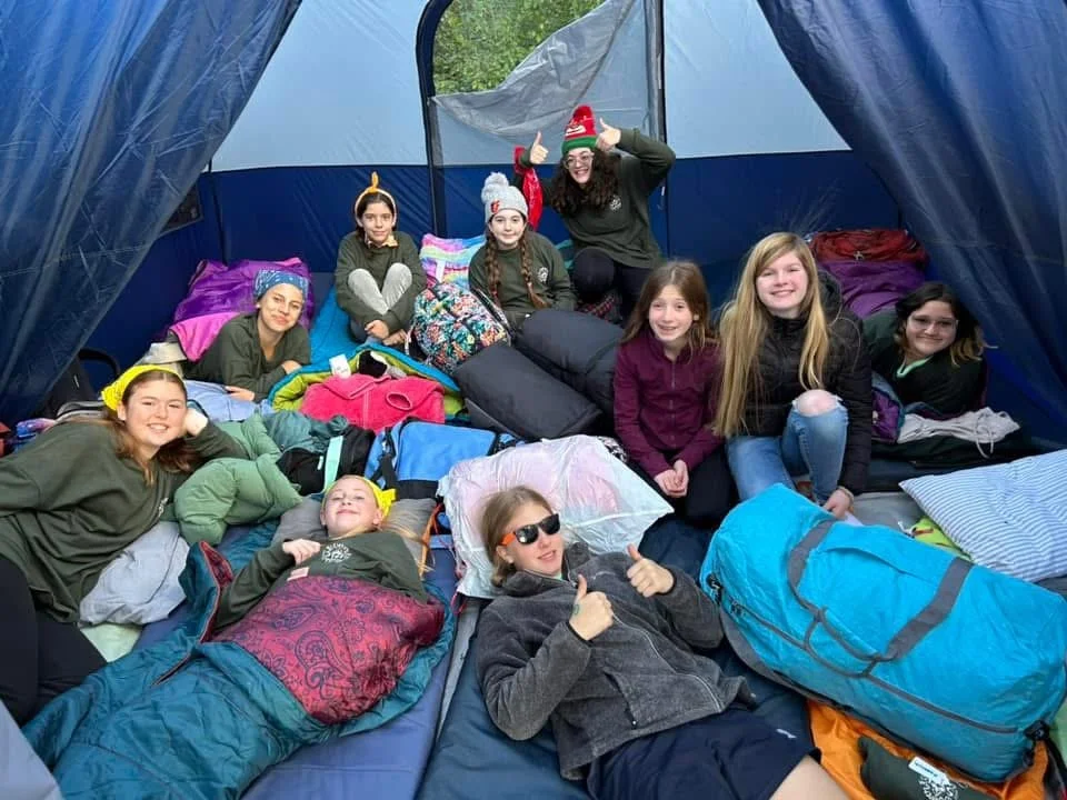 A group of children and a woman inside a camping tent. The children are sitting or lying on sleeping bags and pillows, smiling and enjoying their camping experience. The woman is giving a thumbs-up sign and smiling.
