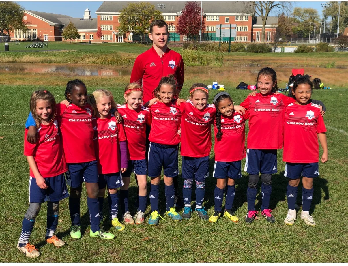 A group of young girls in red soccer uniforms with the logo 'Chicago Edge' and their coach standing on a grassy field, posing for a team photo on a sunny day with trees and a large building in the background.
