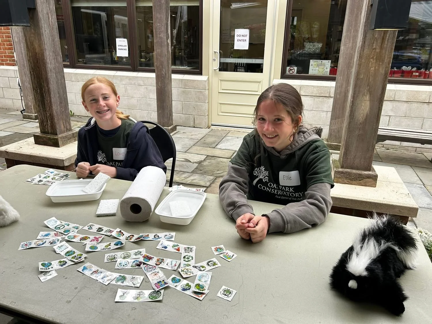 Two smiling teens girls are seated wearing dark green t-shirts that say "OAK PARK CONSERVATORY" at a table outdoors with stickers and a stuffed black-and-white puppy on the table. The background shows a building with large windows and a door.