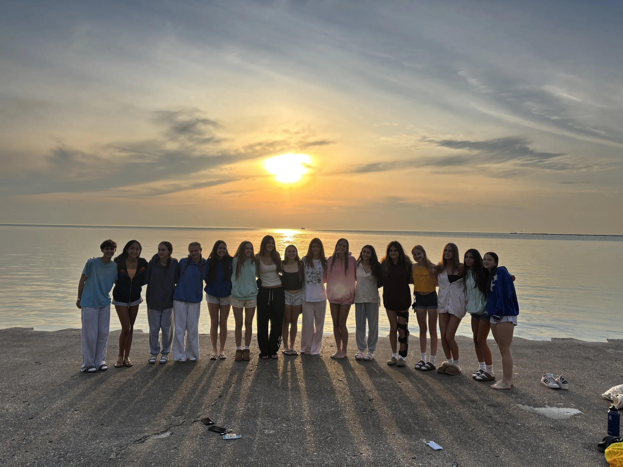 Group of young women standing side by side on a beach at sunset with calm water in the background.