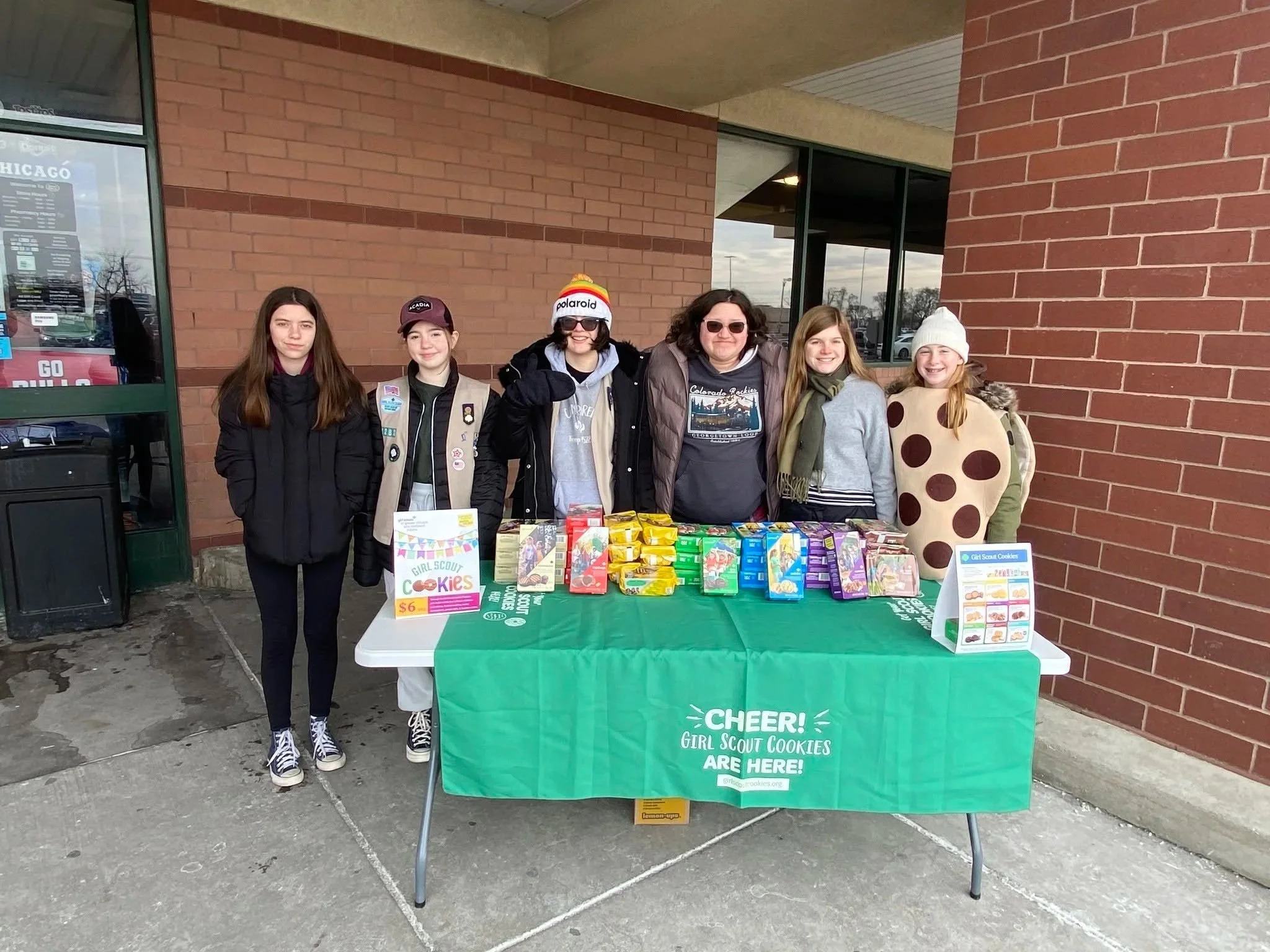 Six teen Girl Scouts standing behind a table with Girl Scout cookies outside a building with brick walls and large windows. The table is covered with a green tablecloth that reads 'CHEER! GIRL SCOUT COOKIES ARE HERE!'