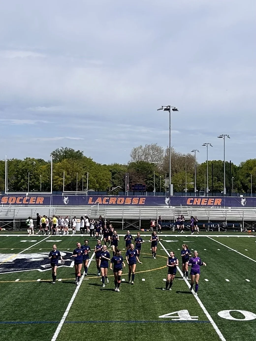 Girls' lacrosse team walking on a sports field with bleachers in the background, under a partly cloudy sky.