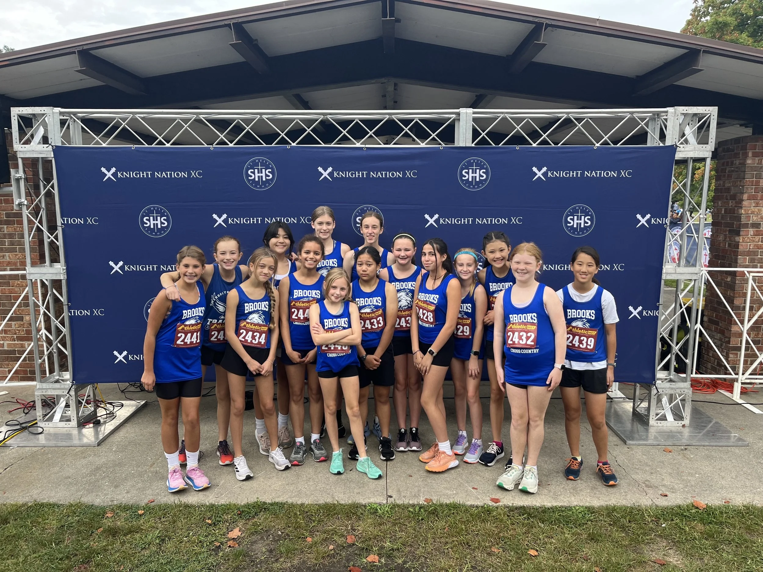 Group of young female cross-country runners in blue uniforms standing together in front of a blue backdrop with 'KNIGHT NATION XC' and 'SHS' logos at an outdoor event.