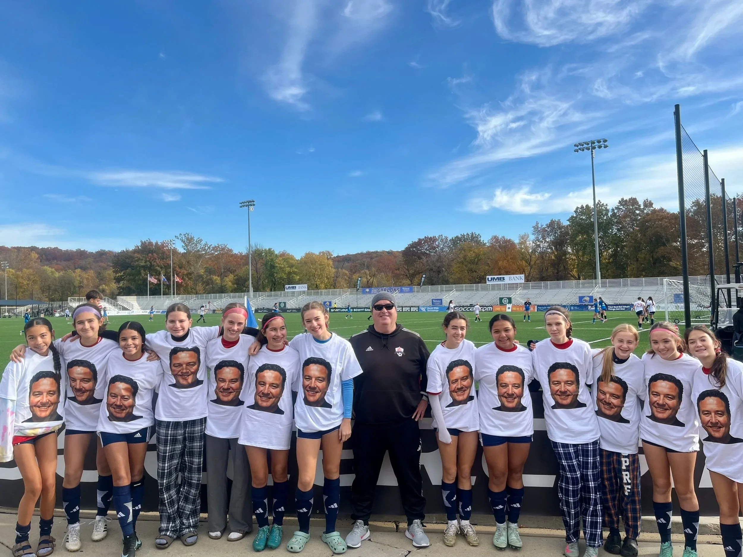 A group of young female soccer players and their coach smiling for a photo on the field after a game, with a stadium, trees, and a blue sky in the background.