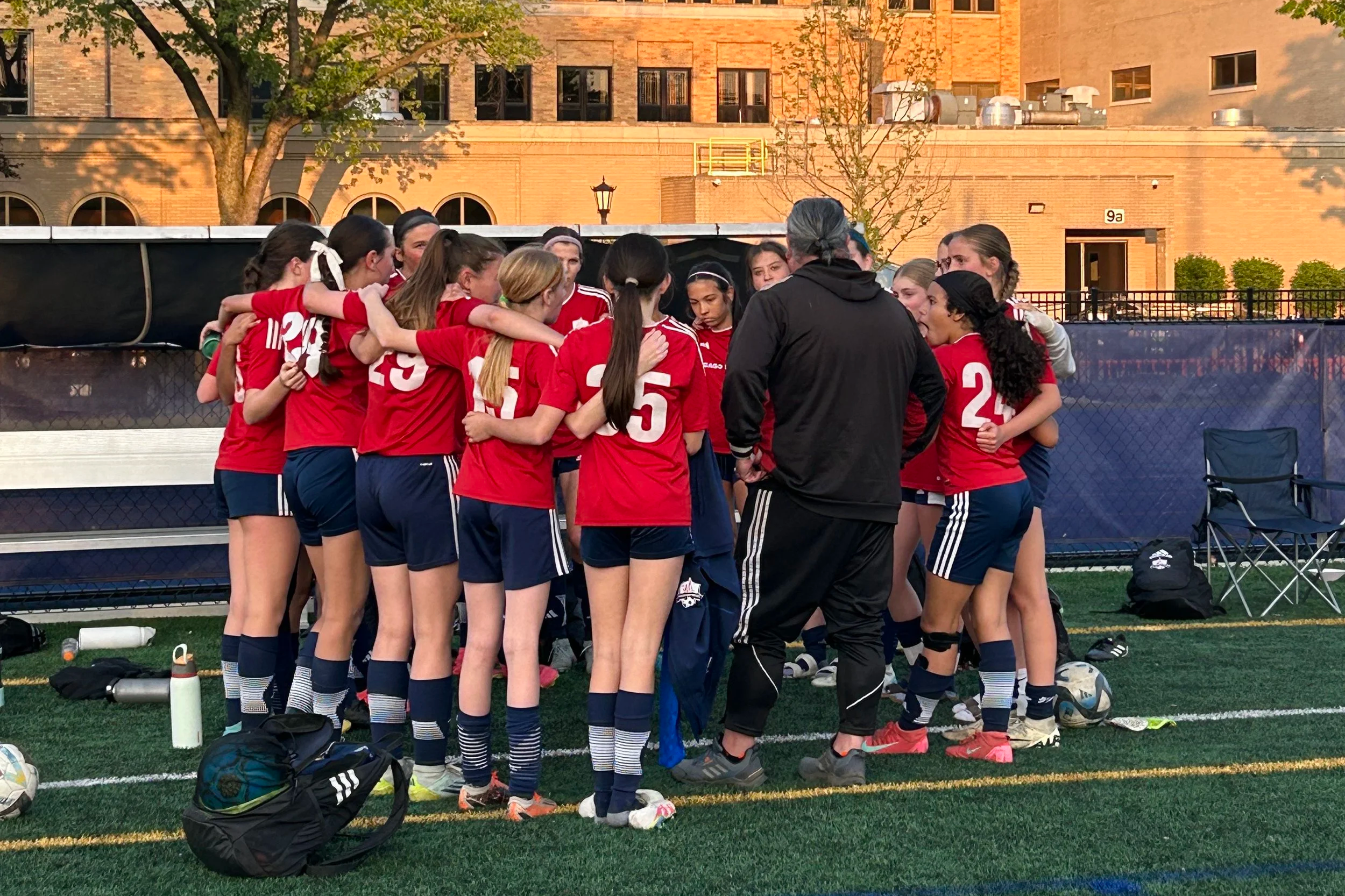 A group of young female Chicago Edge soccer players wearing red jerseys, dark shorts, and knee-high socks gathered around their coach on a soccer field during sunset, with equipment and water bottles on the ground.