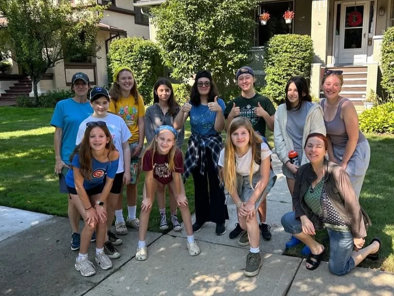 Group of ten teen Girl Scouts and two adults standing and kneeling on a sidewalk in front of a house with green bushes and trees, all smiling and posing for the photo.
