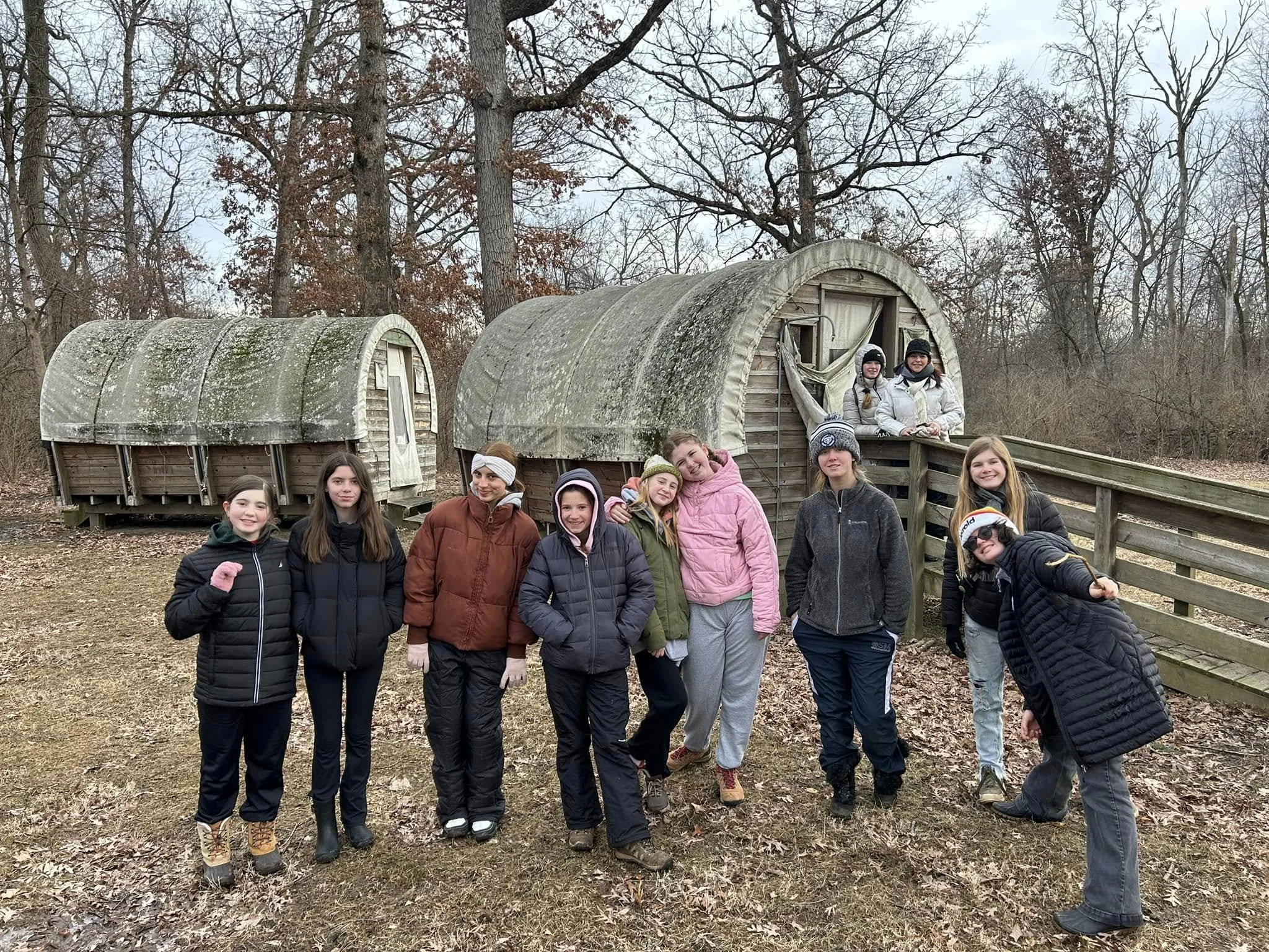A group of young teen girls wearing winter jackets and hats, posed outdoors in front of two covered wagons with curved roofs, surrounded by leafless trees on a cloudy day.