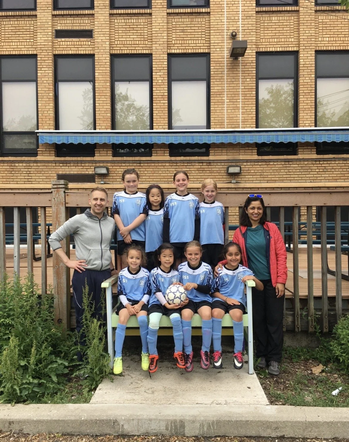 A youth girls' soccer team with two coaches or teachers, posing for a group photo outside in front of a brick building. The team is wearing blue and black soccer uniforms, and one girl in the front row is holding a soccer ball. The girls are smiling, and the photo appears to be taken on a cloudy day.