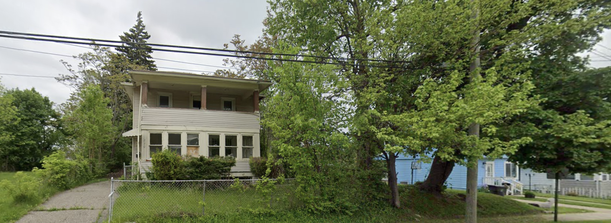 A two-story house with a front porch, surrounded by green trees and a chain-link fence in a residential neighborhood.