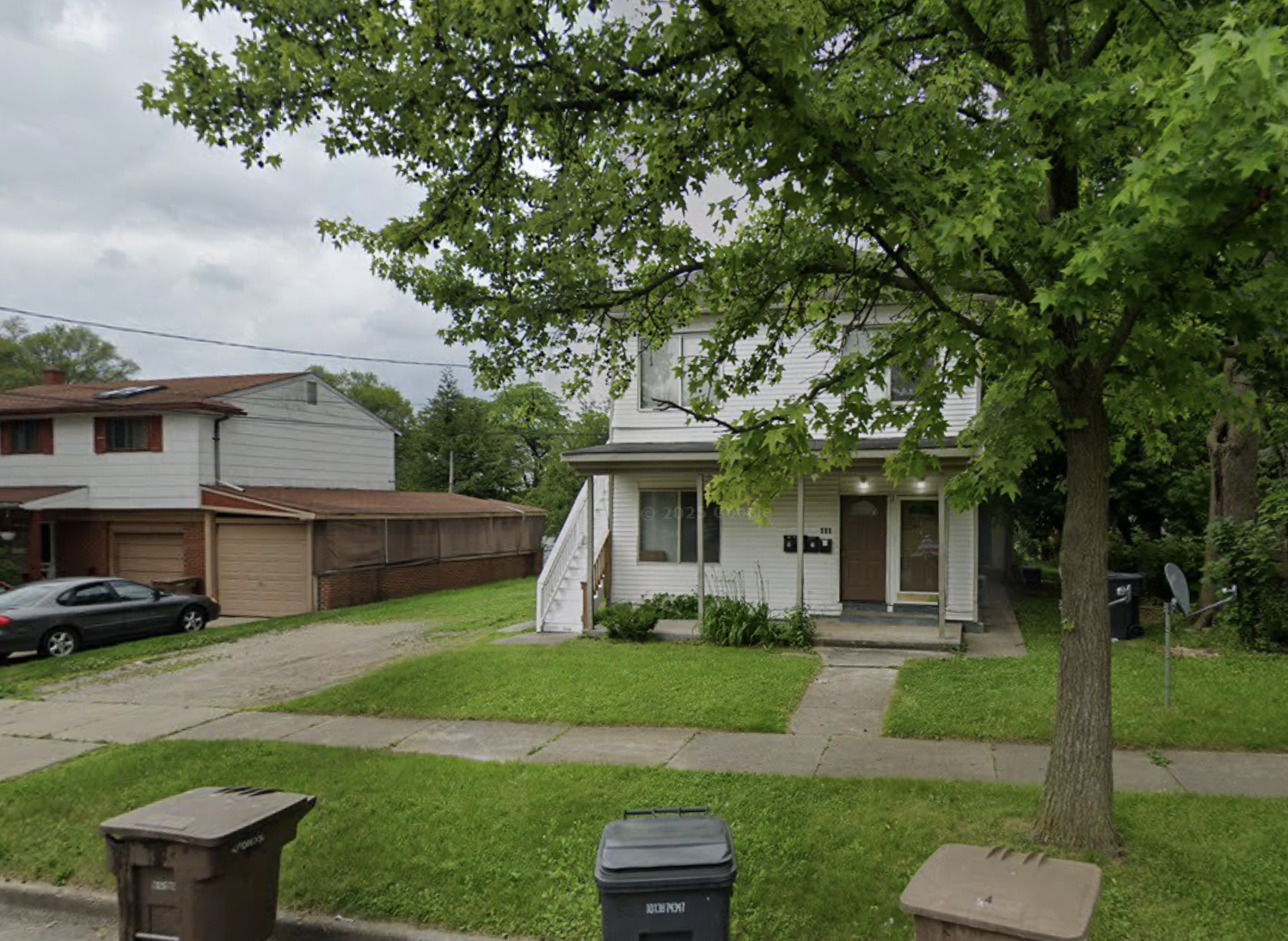 A two-story white house with brown front door and stairs, large tree in front, sidewalk, mailbox, trash bins, parked cars, neighboring house, cloudy sky.