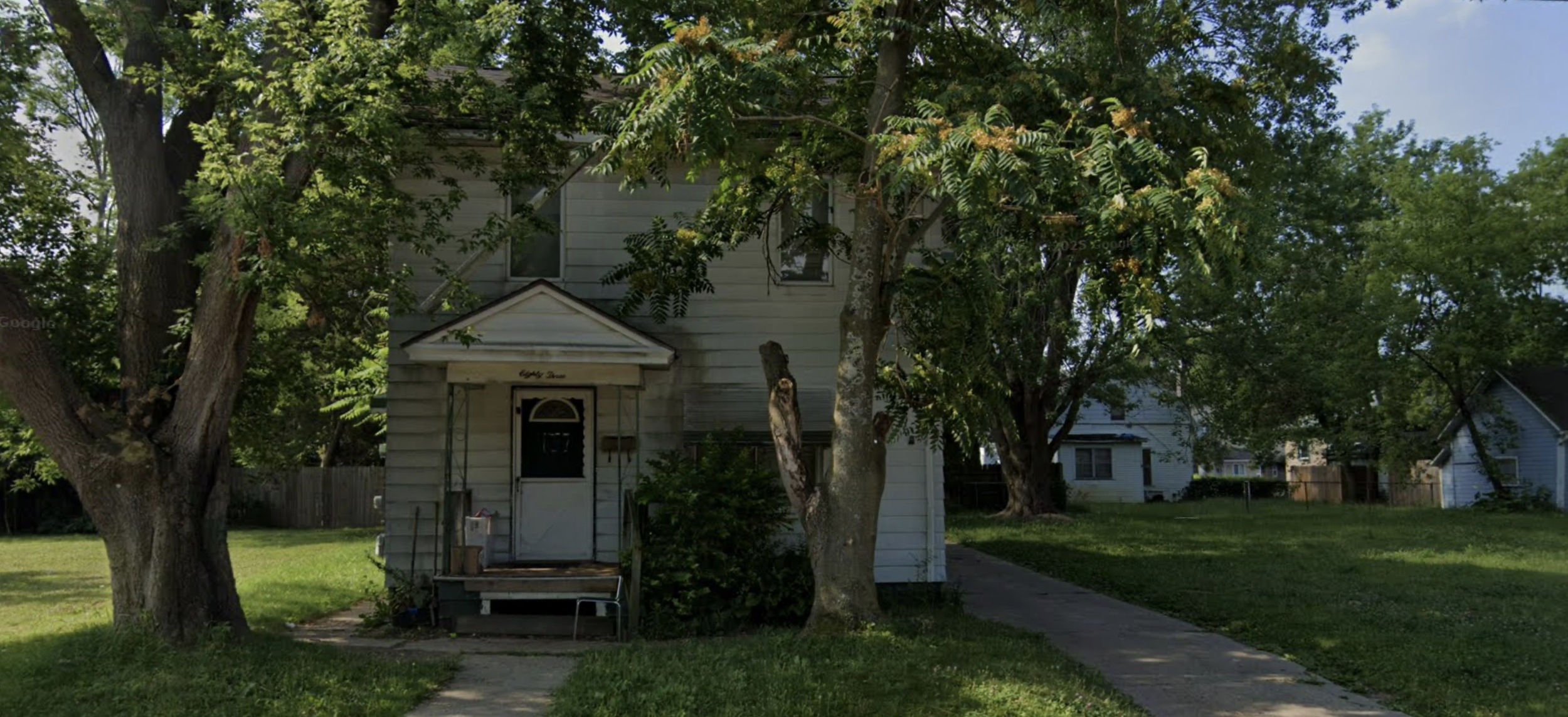 Small white house with a front porch, surrounded by tall trees and green grass in a suburban neighborhood.