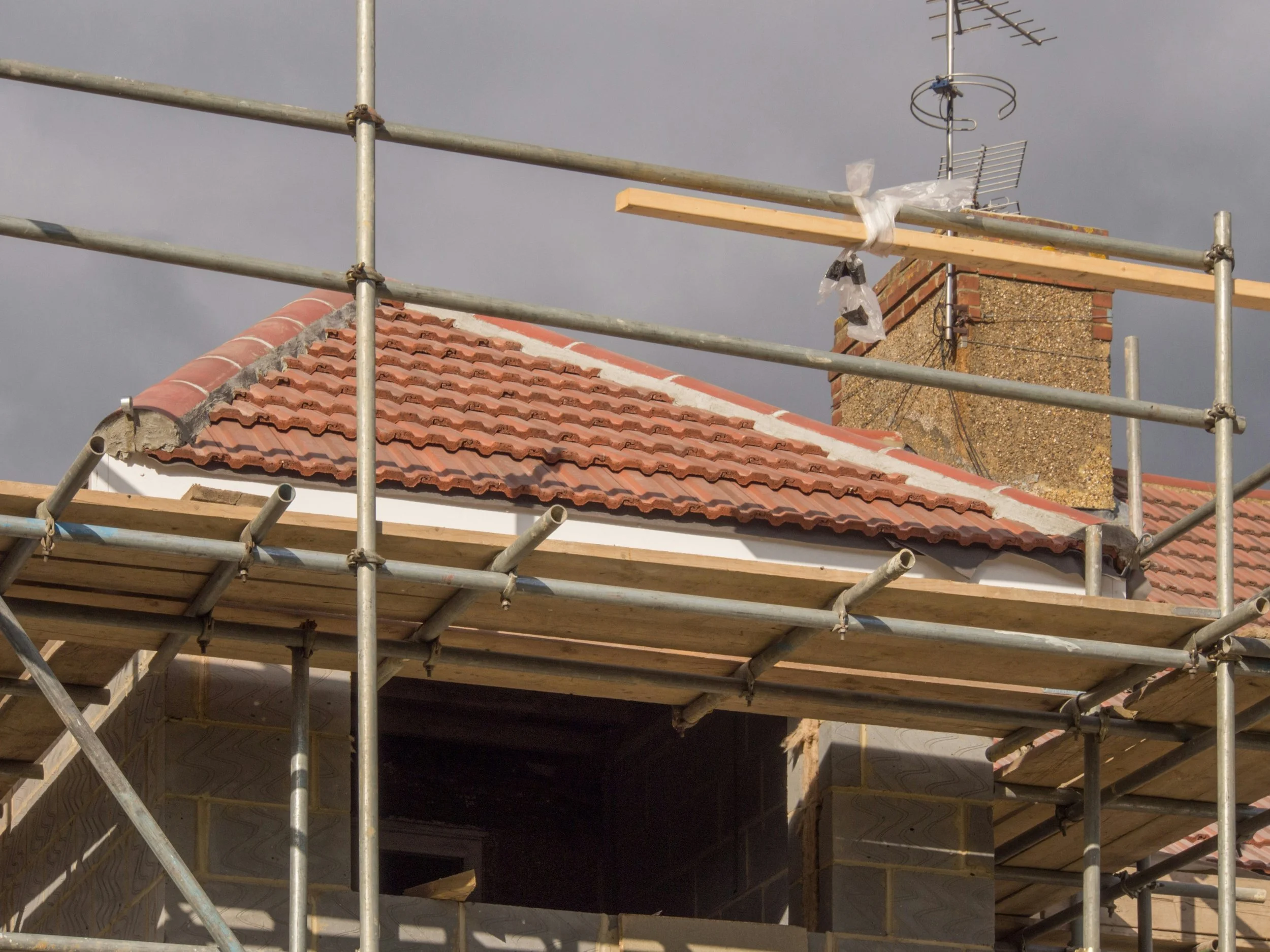 Construction scaffolding around a house with a red tile roof and a brick chimney, with antennas visible on top of the chimney, against a cloudy sky.