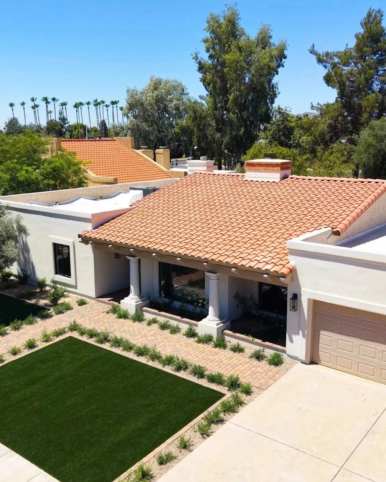 A modern house with a red tile roof, white exterior walls, a small front porch with columns, a driveway, and a lawn with a paved walkway and plants, set in a sunny suburban neighborhood with trees and other houses in the background.