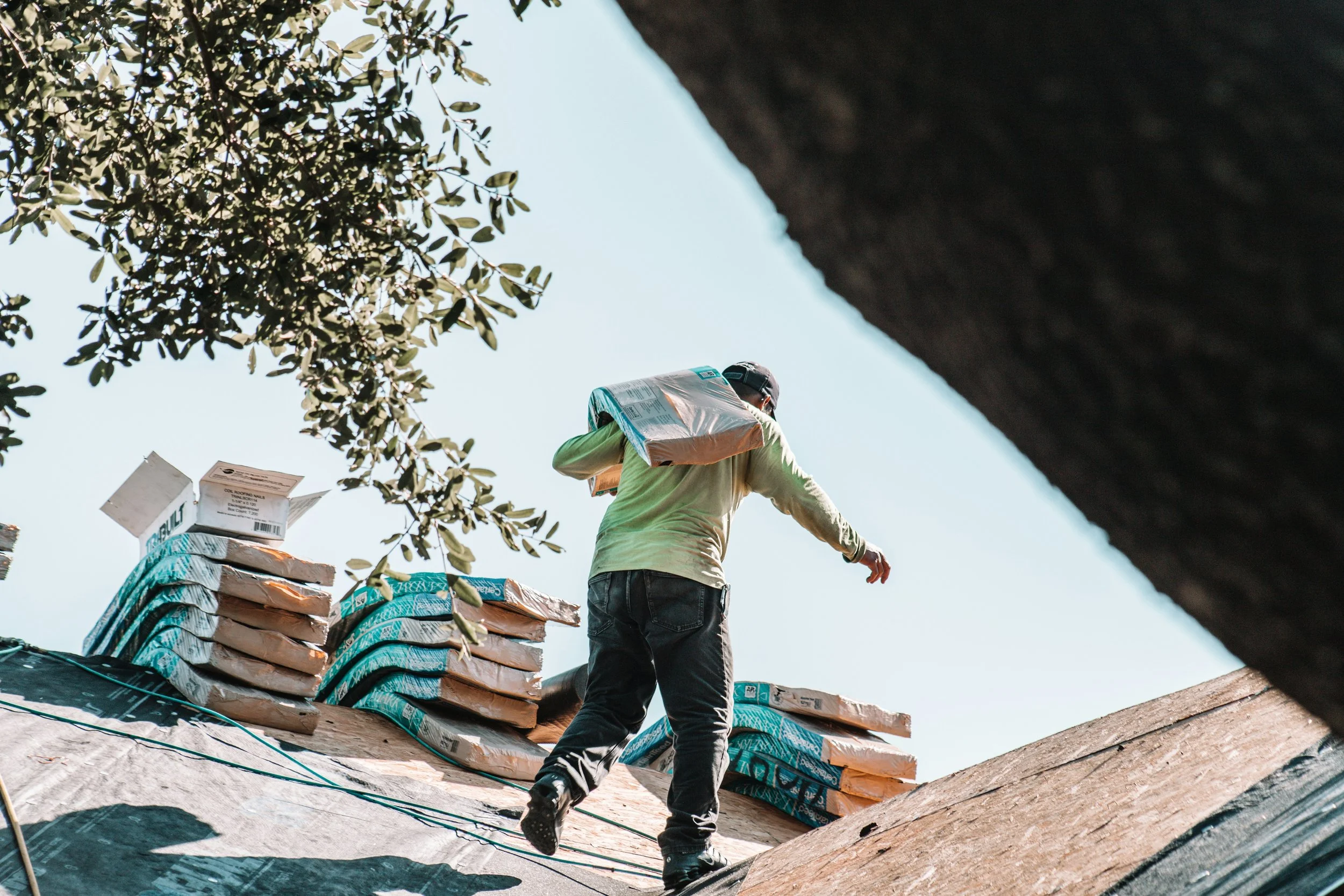 A construction worker wearing a green shirt and black cap walking on a sloped roof surrounded by stacks of roofing materials. The scene is outdoors with sky and tree branches visible.