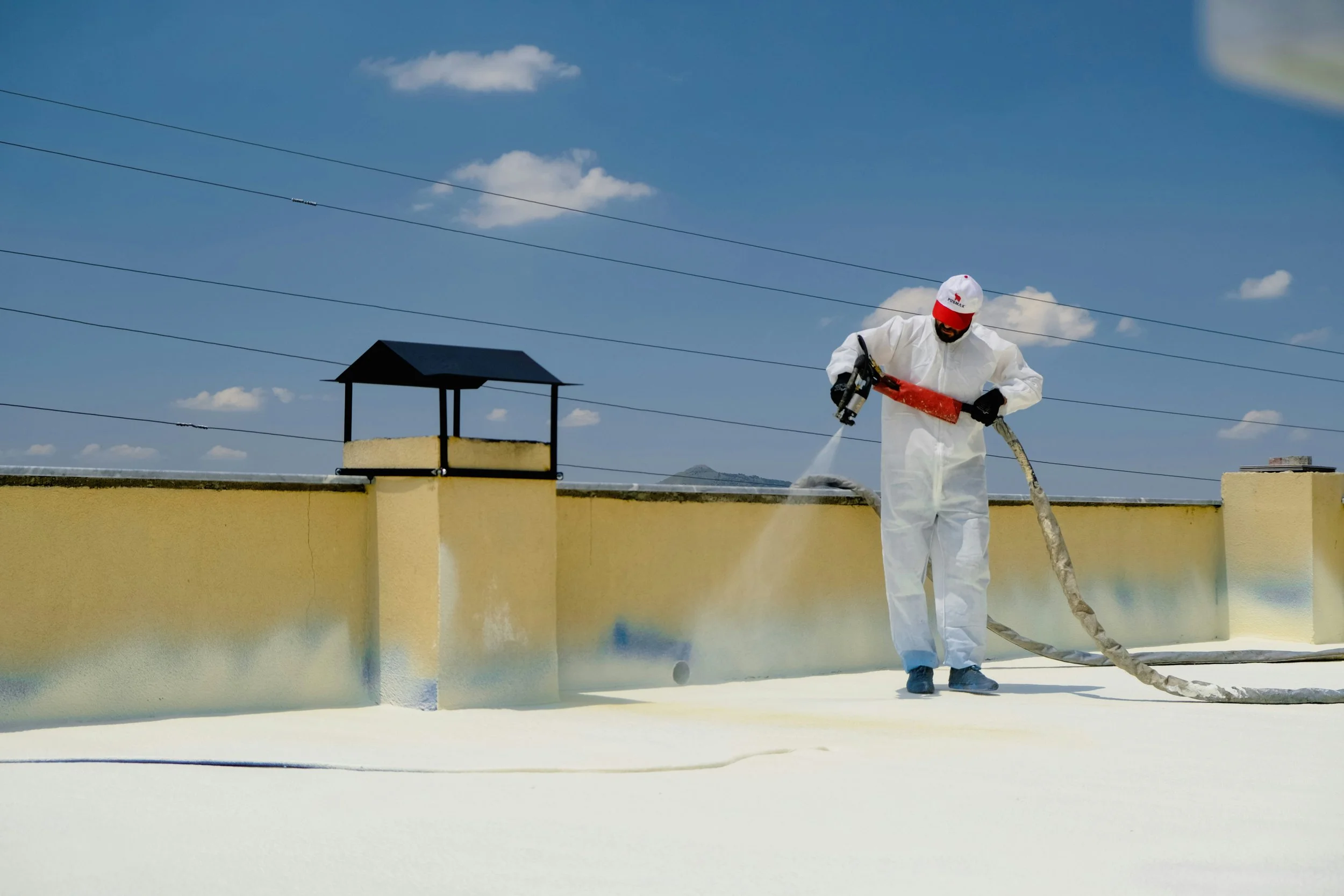 Person in white protective suit and red cap cleaning a roof with a hose against a blue sky with clouds.