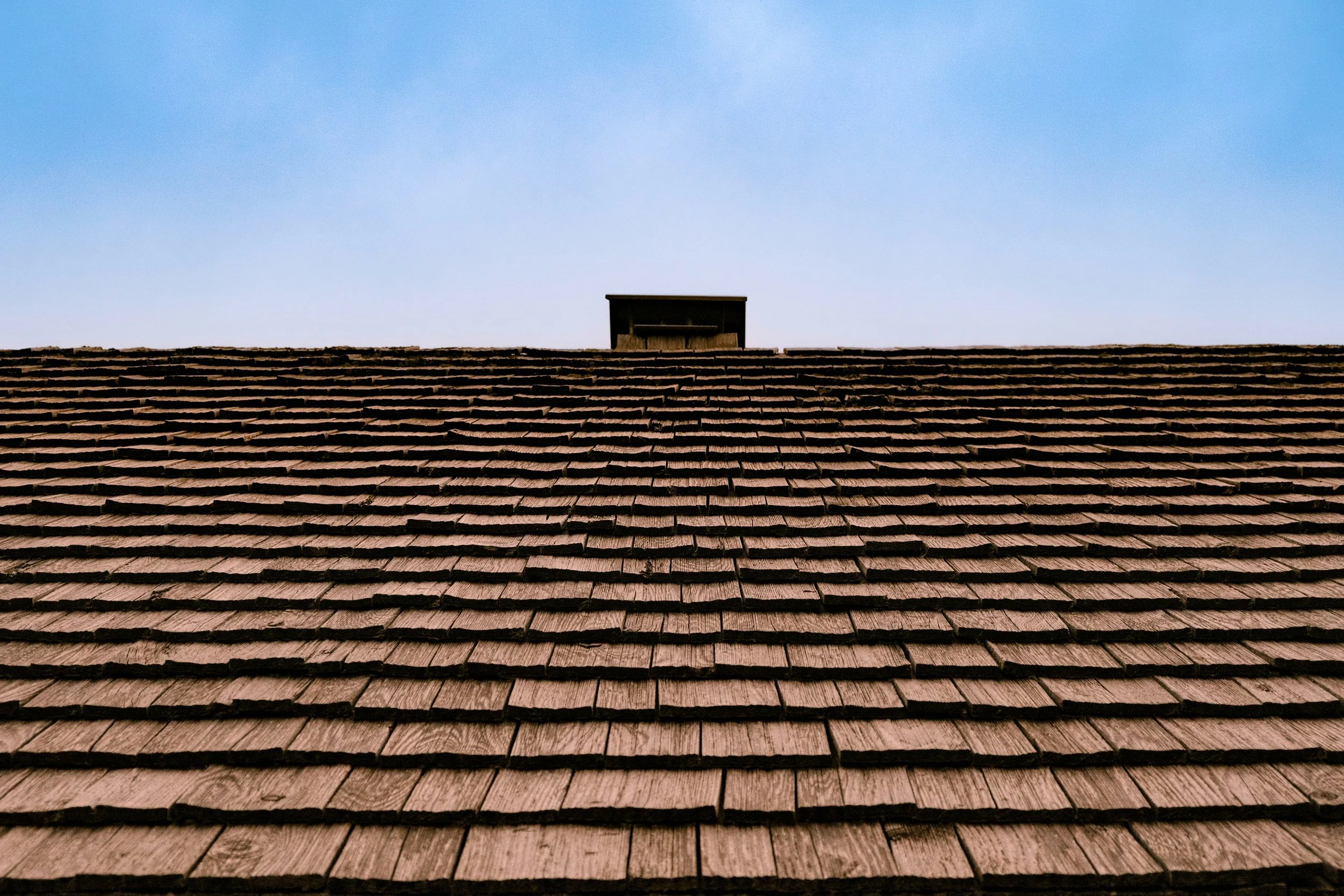 A rooftop with brown shingles and a small structure against a bright blue sky.