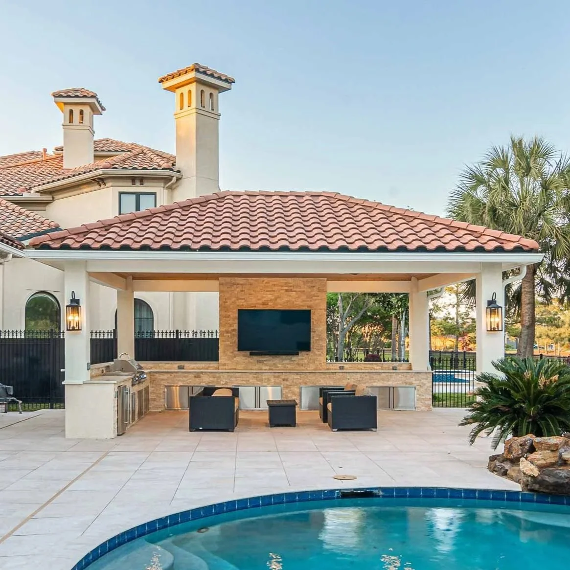 Poolside outdoor living area with a brick fireplace, mounted TV, seating, and built-in grill, with a house featuring tile roofs and palm trees in the background.