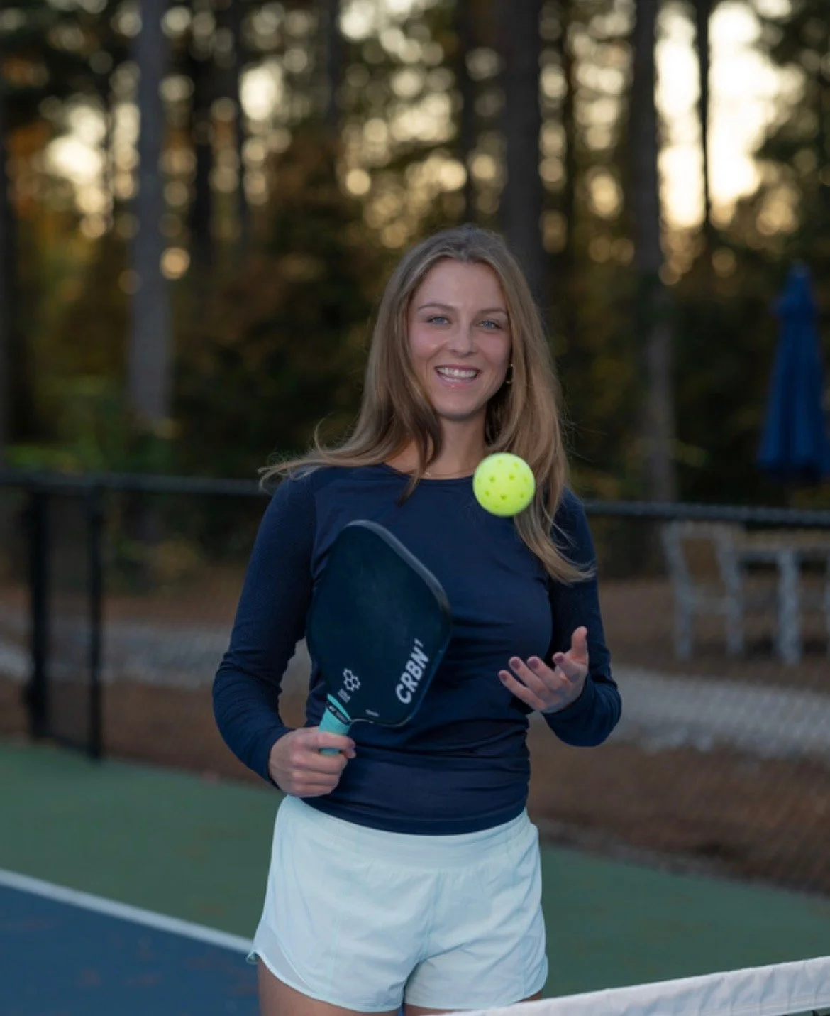 A woman standing on a tennis court holding a paddle and a tennis ball, smiling at the camera with trees in the background.