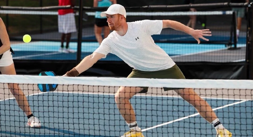 A man playing pickleball on an outdoor court, reaching to hit a yellow ball with a paddle.