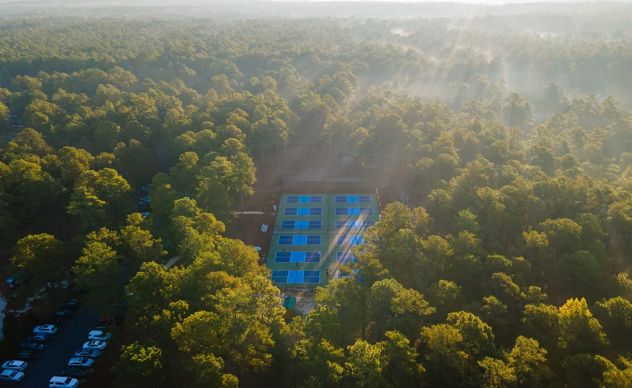 Aerial view of a densely wooded forest with a tennis court equipped with solar panels in the center, and a parking lot visible at the bottom left, with morning sunlight streaming through mist.