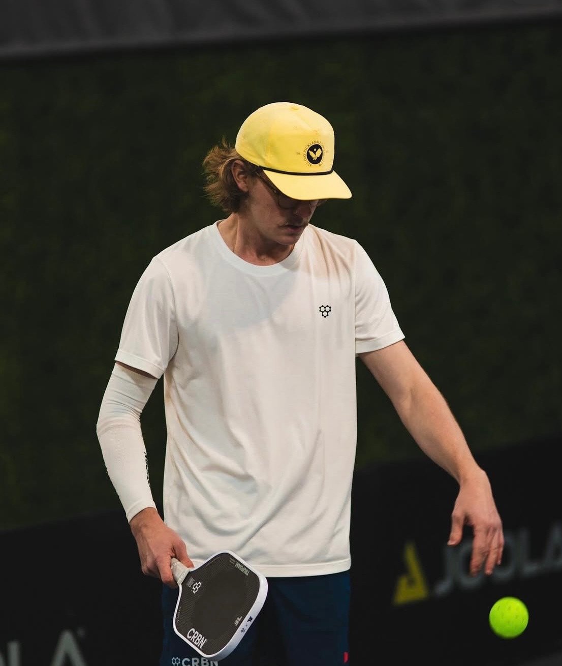 A man wearing a yellow cap and white t-shirt stands on a tennis court holding a paddle, with a tennis ball nearby.
