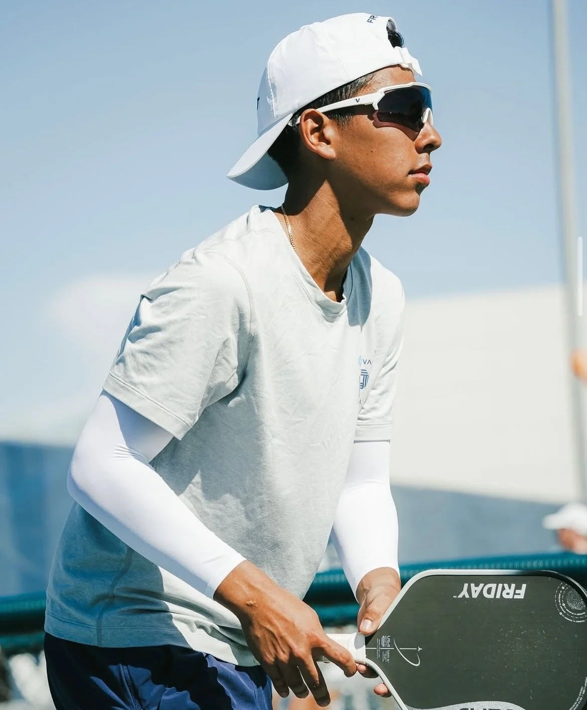 A young man in athletic gear with sunglasses, a white cap worn backwards, and a white long-sleeve shirt underneath a short-sleeve shirt, holding a paddle in a beach or by-water setting.