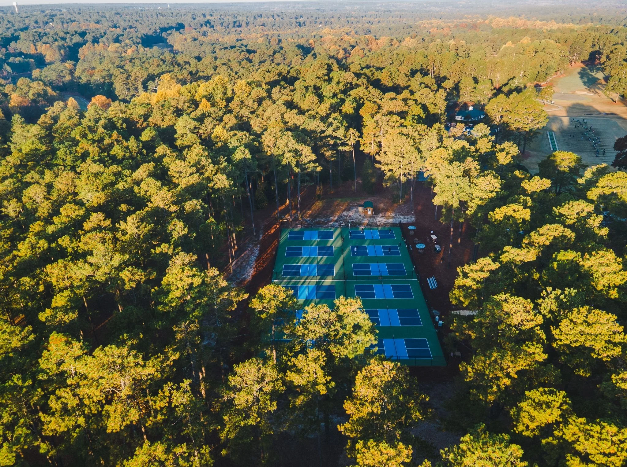 An aerial view of a wooded area with dense trees in fall colors, a parking lot, and multiple tennis courts.