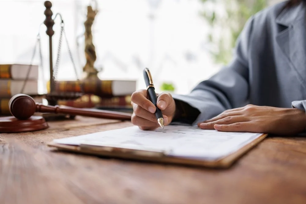 Person in a suit signing a document at a wooden desk, with legal books, a gavel, and scales of justice in the background.