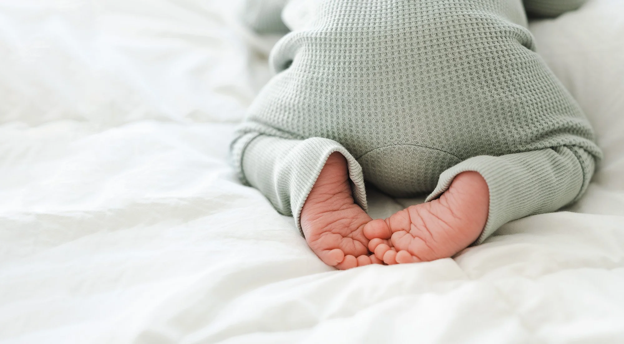 Close-up of a baby laying on a white bed, wearing light gray textured pajamas.