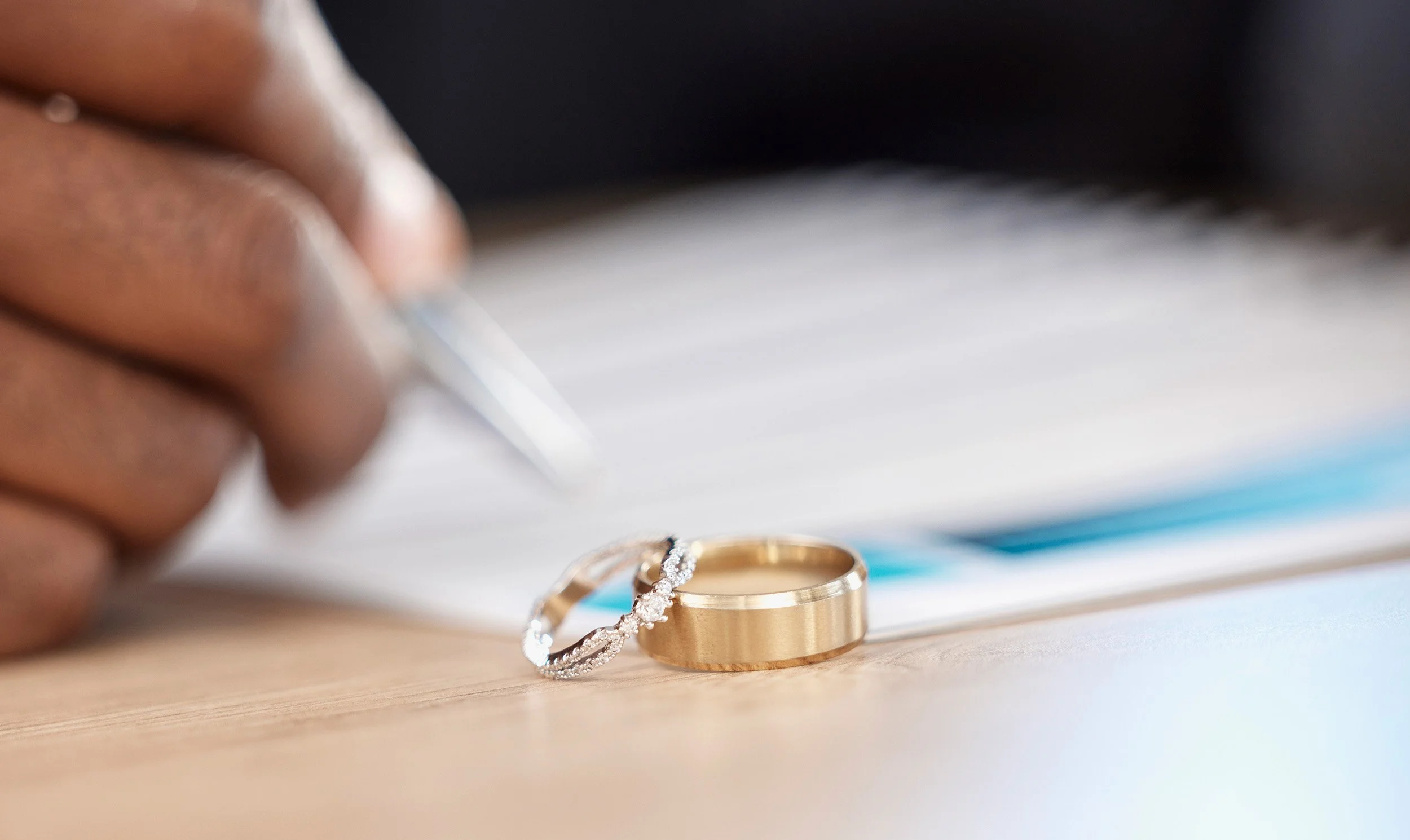 Close-up of a hand holding a pen near a gold wedding band and an engagement ring with diamonds, placed on a wooden surface with a blurred background of open book pages.