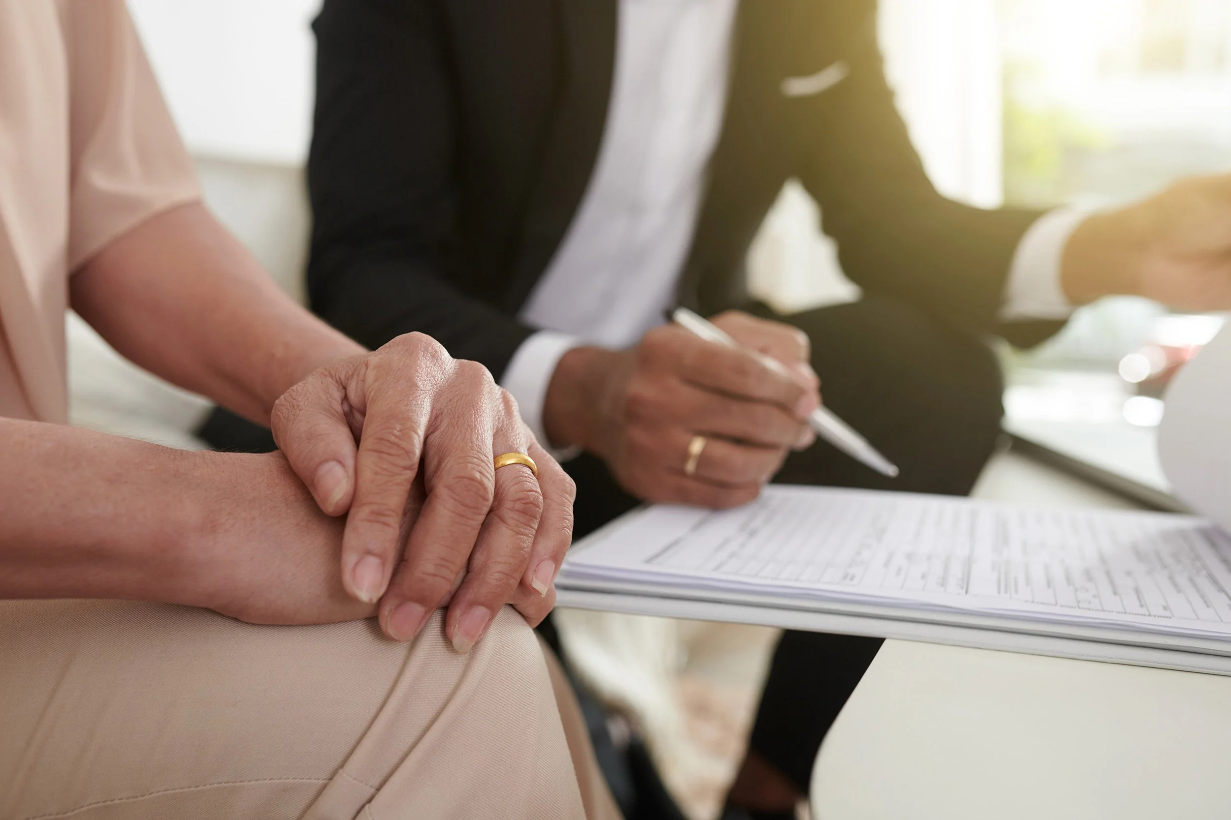 Close-up of hands, one with a wedding ring, resting on a person's arm during a formal meeting or discussion. Other individuals are taking notes or signing documents at a table with a notebook and pen, with sunlight coming through a window in the background.