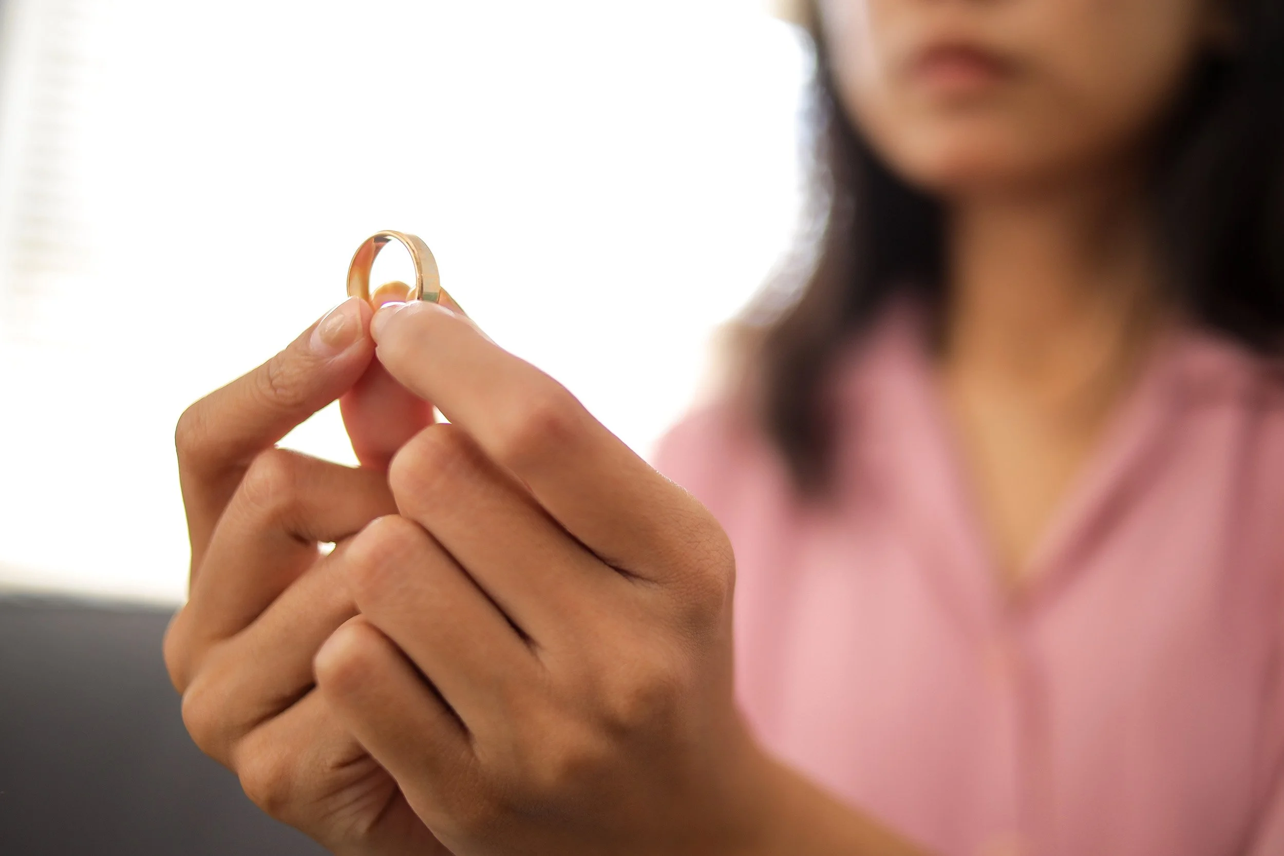 A woman holding a gold wedding ring between her thumb and index finger, with her face blurred in the background.