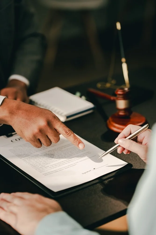 A person is pointing at a signed legal document on a table while another person holds a pen, with courtroom gavel, notepad, and scale visible in the background.