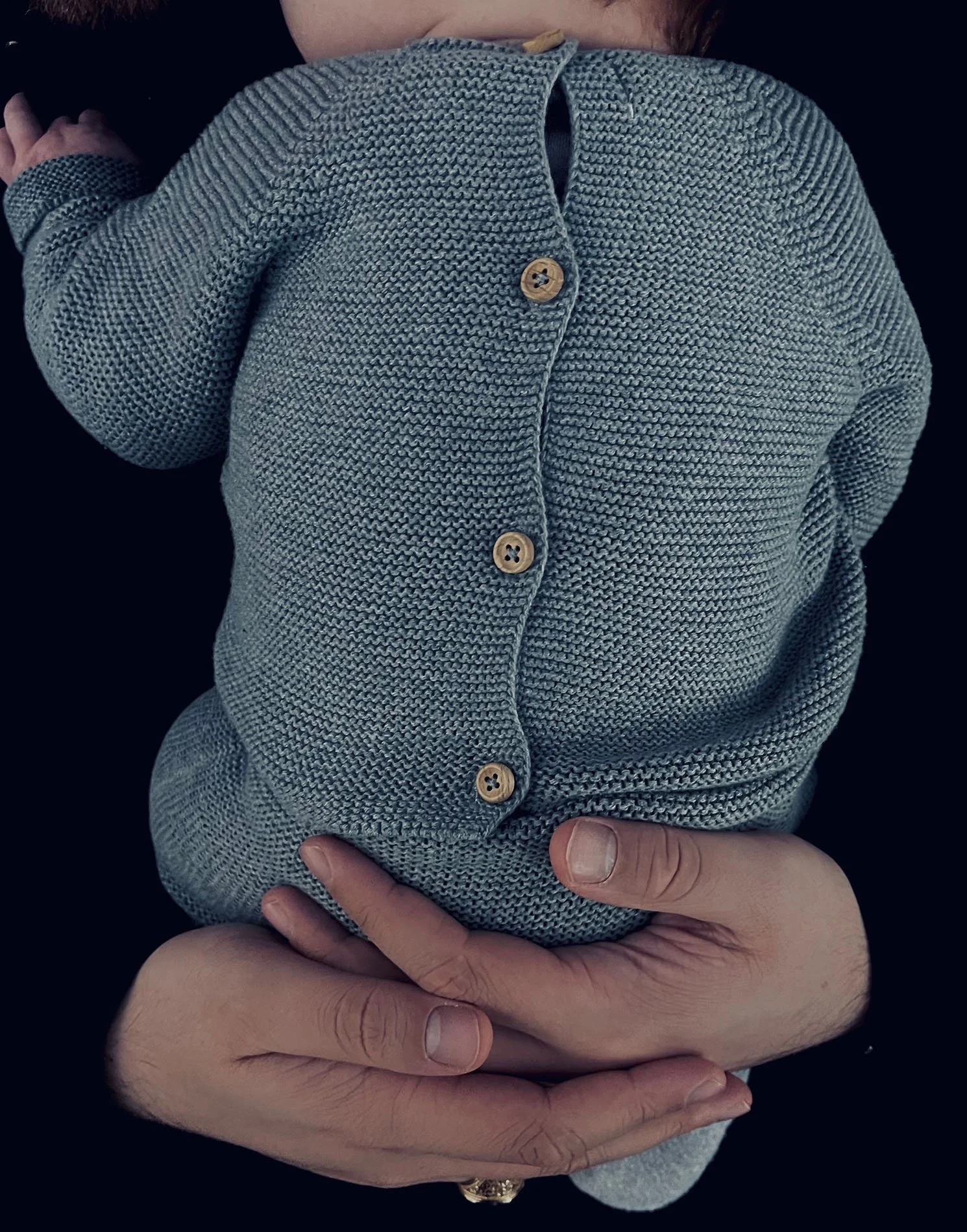 A person holding a baby dressed in a gray knit sweater with wooden buttons, lying on their chest against a black background.
