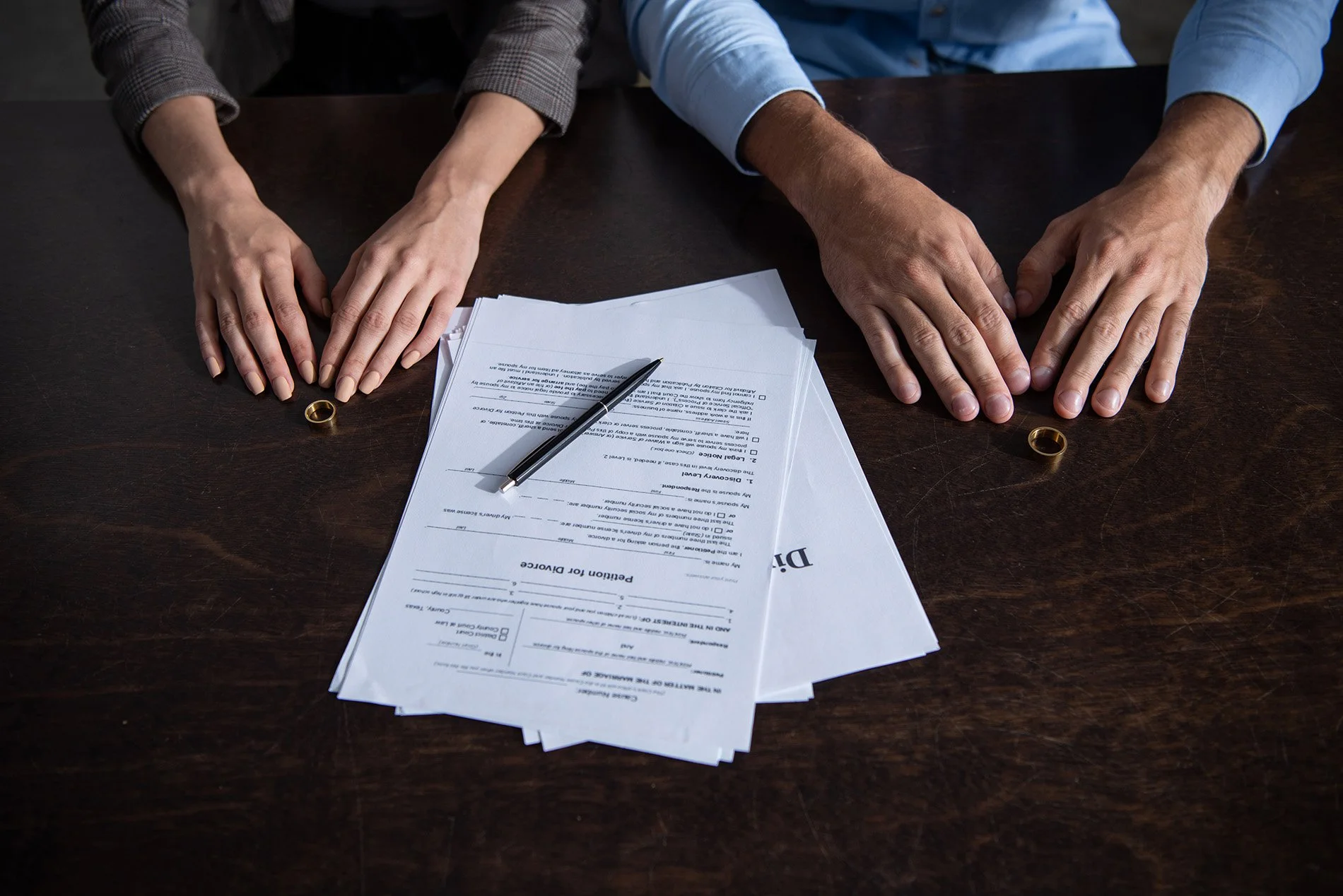 Two people sitting at a dark wooden table, with one hand of each person resting on the table near wedding rings. In front of them are several insurance claim forms, a pen on top of the forms, and a label that reads 'Claim'.