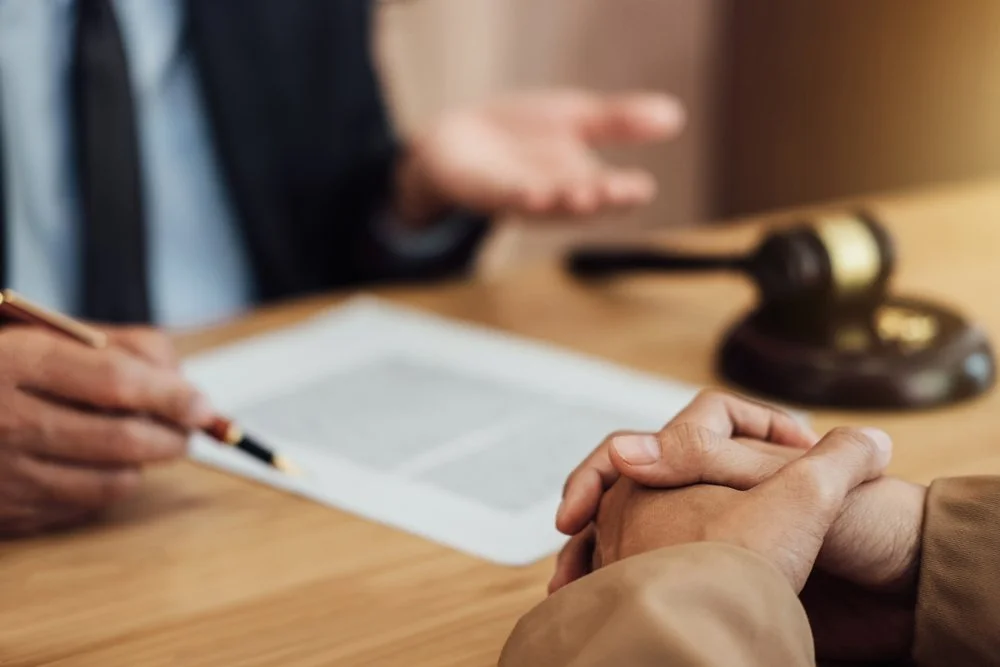 Two people sitting at a table during a legal or formal meeting, with one person holding the other's hands, a gavel and document on the table, and a blurred figure in the background gesturing with their hand.