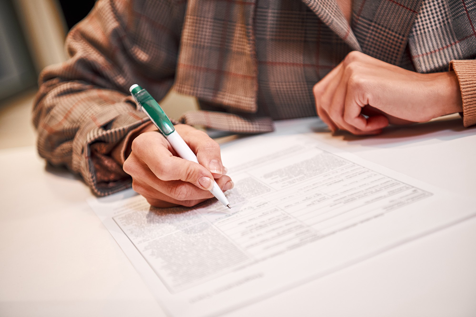 A person wearing a plaid shirt signing a document with a pen on a table.