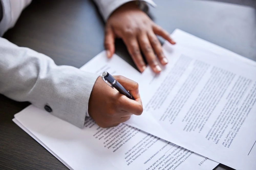 A person in a grey suit is signing a document with a black pen, while their other hand rests on the paper on a dark wooden table.