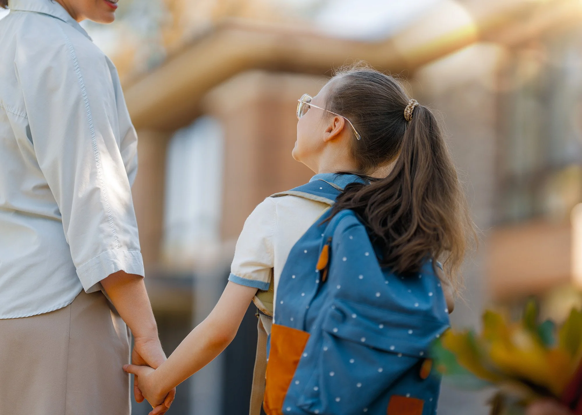 A young girl with glasses and a ponytail holding hands with an adult, possibly her mother, in an outdoor setting with trees in the background.