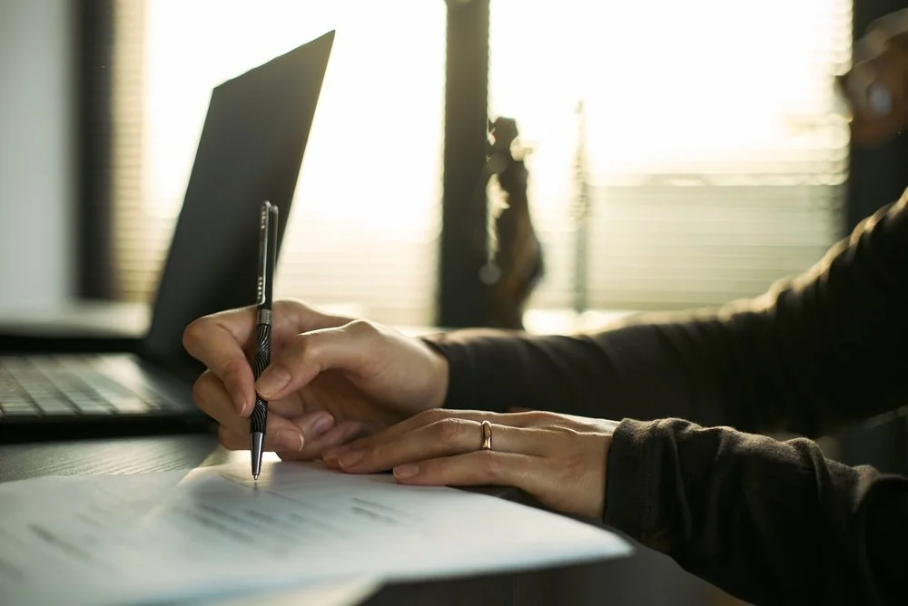 A person writing on a paper with a pen on a desk, with a laptop and a small statue in the background.