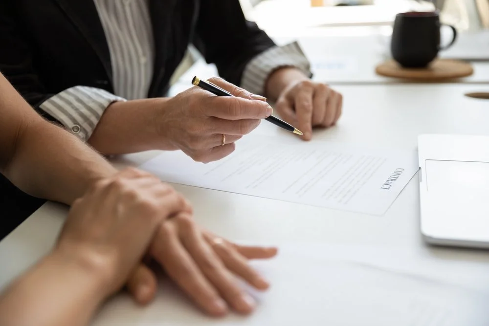 Two people reviewing and signing a document at a table in an office, with a coffee mug and a laptop nearby.