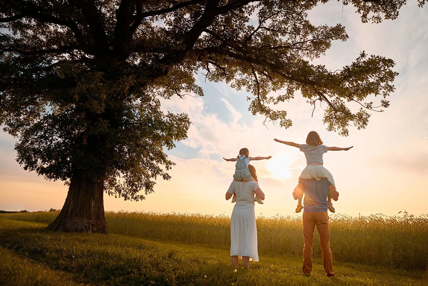 Family of four outdoors at sunset with large tree; two adults and two children, one girl on adult's shoulders, both with arms outstretched.