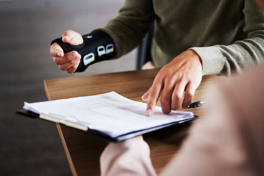 Person wearing a wrist brace and pointing at documents on a desk with another person, with one hand on the documents.