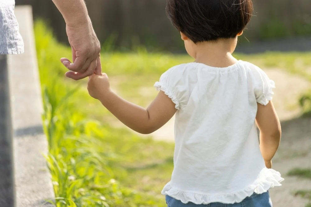 A young girl holding hands with an adult while walking outdoors in a grassy area.